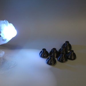 May include: A bright blue light bulb with smoke coming from it, illuminating a group of seven black plastic objects on a white surface.