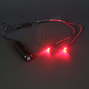 Op de afbeelding: Een zwart elektronisch apparaat met twee rode LED-lampjes. Het apparaat heeft een zwart snoer en twee rode draden die naar de lampjes lopen. De lampjes zijn verlicht en geven een heldere rode gloed af.