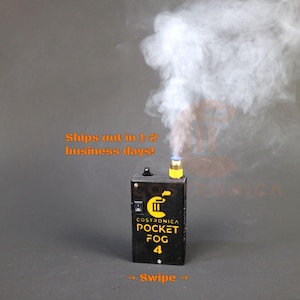 Op de afbeelding: Een zwarte Costronica Pocket Fog 4 machine die witte rook uitstoot tegen een grijze achtergrond. De machine heeft gele en blauwe accenten. De tekst "Pocket Fog 4" en "Swipe" zijn zichtbaar op het apparaat.