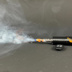 Puede incluir: Un dispositivo negro con el texto "COSTSONIC" y un logotipo, que emite una nube de vapor blanco. El dispositivo tiene detalles en naranja y azul, y un pequeño mando a distancia está al lado.