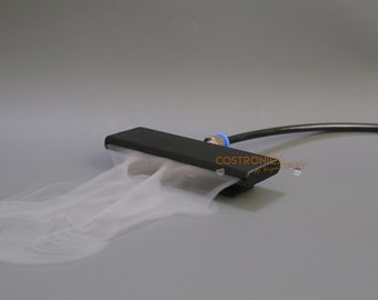 May include: A black rectangular device with a nozzle, emitting a stream of white vapour. A black tube and blue fitting are attached. The device is on a grey surface. The text "COSTRONICA" is visible on the device.