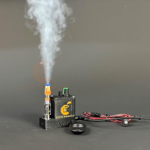 Puede incluir: Un dispositivo negro con el texto "COSTRONICA" y un logotipo, que emite una columna de vapor blanco. El dispositivo tiene una perilla verde y está conectado a un mando a distancia y cables. El vapor se eleva desde un atomizador transparente y naranja.