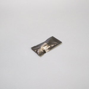 Peut inclure: Un petit sachet rectangulaire en plastique noir semi-transparent contenant un objet métallique. L'emballage est scellé et présente une surface sombre légèrement réfléchissante qui masque partiellement le contenu, révélant une forme métallique.