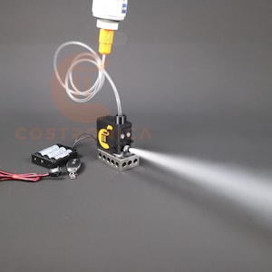 Könnte beinhalten: Eine kleine schwarze und silberne Nebelmaschine mit einem durchsichtigen Schlauch, der mit einer gelben und weißen Flasche verbunden ist. Die Maschine erzeugt einen weißen Nebel. Die Maschine ist mit einem schwarzen Akkupack und einer Fernbedienung verbunden.