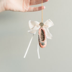 Puede incluir: Un adorno de zapato de ballet rosa con purpurina dorada y detalles de cinta blanca. El adorno cuelga de una cuerda dorada.