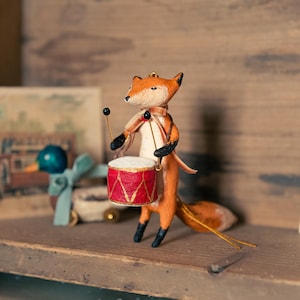 Puede incluir: Una pequeña figura de zorro naranja tocando un tambor rojo con detalles dorados. El zorro sostiene dos baquetas y lleva una bufanda negra y blanca alrededor del cuello. La figura está en un estante de madera con una postal vintage en el fondo.