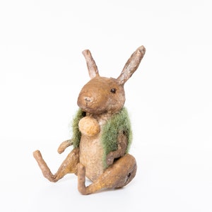 May include: A brown and green handmade bunny rabbit figurine with long ears. The bunny is sitting with its legs crossed and holding a small brown ball in its paws. The bunny is wearing a green moss-like jacket.