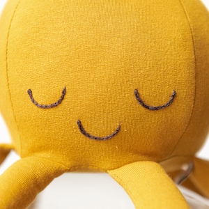 May include: A yellow plush octopus with a smiling face. The octopus has eight tentacles and is made of soft fabric.