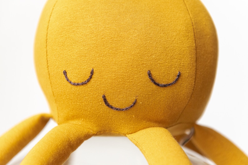 May include: A yellow plush octopus with a smiling face. The octopus has eight tentacles and is made of soft fabric.