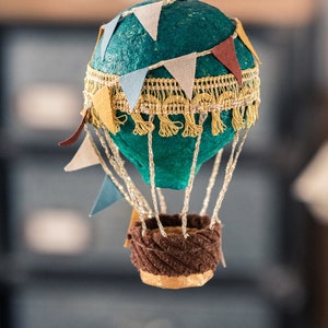 May include: A teal blue hot air balloon ornament with a gold trim and colorful pennants. The balloon has a brown wicker basket.