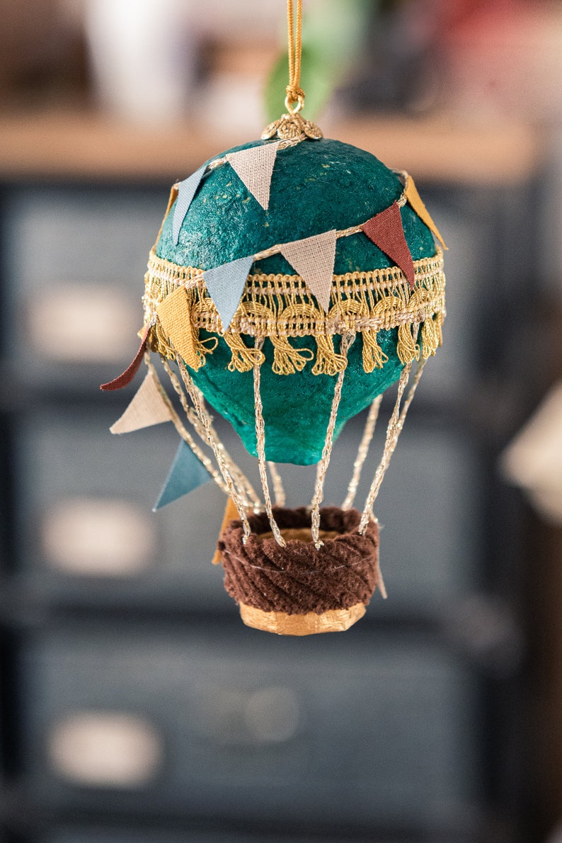 May include: A teal blue hot air balloon ornament with a gold trim and colorful pennants. The balloon has a brown wicker basket.