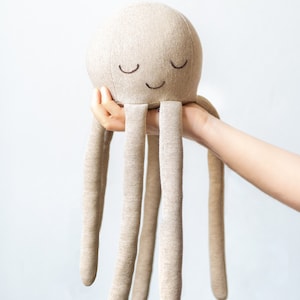 May include: A beige plush octopus toy with a smiling face and closed eyes. The octopus has six long tentacles.