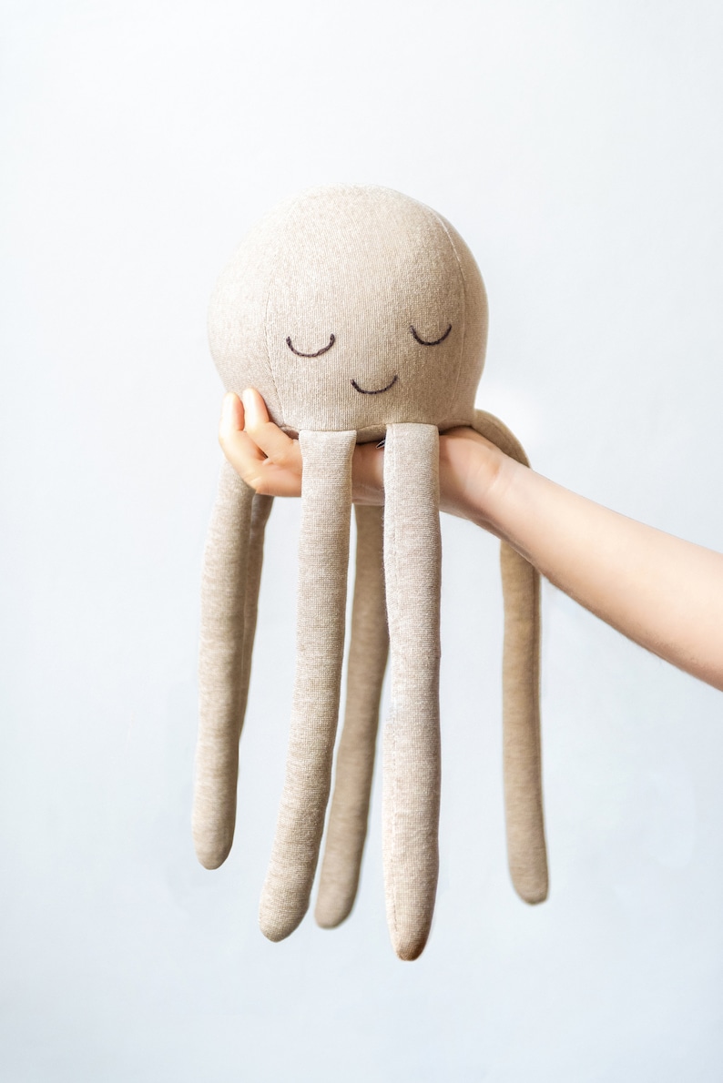 May include: A beige plush octopus toy with a smiling face and closed eyes. The octopus has six long tentacles.