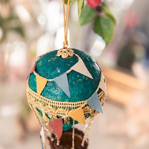 May include: A teal blue hot air balloon ornament with gold trim and colorful pennants. The balloon has a brown wicker basket and is hanging from a gold string.