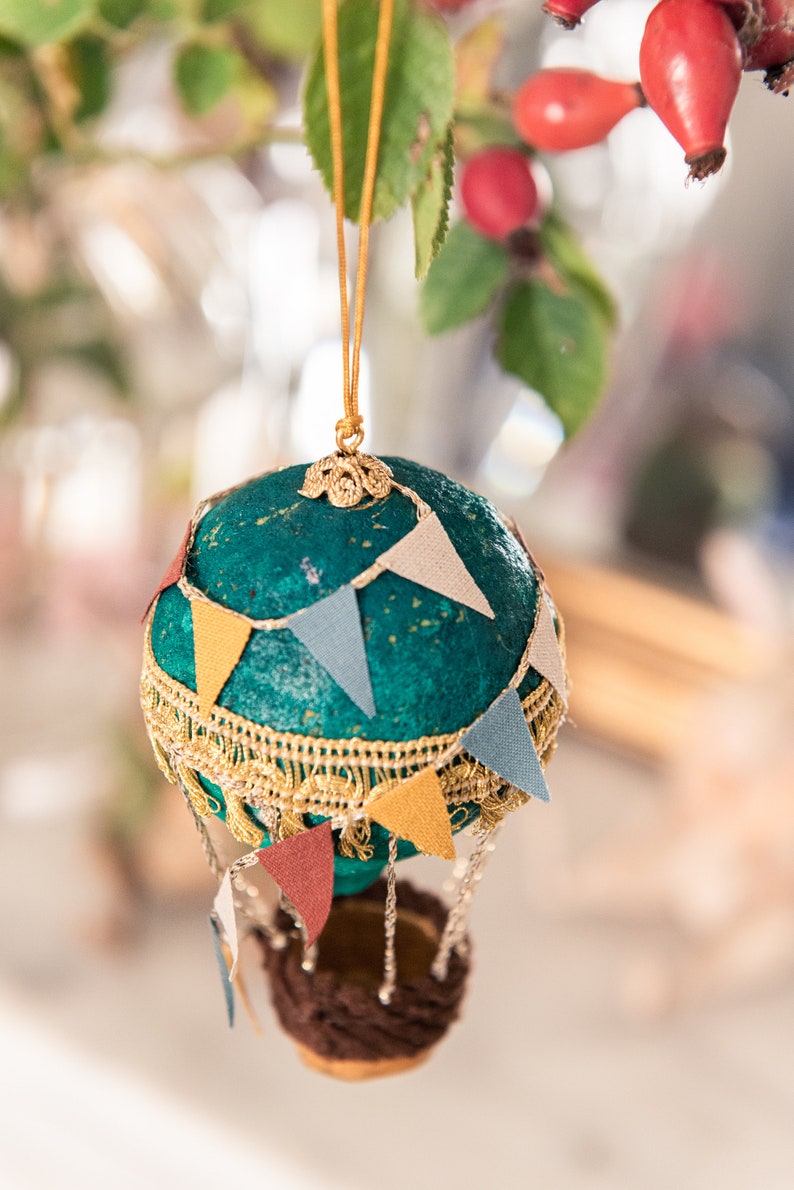 May include: A teal blue hot air balloon ornament with gold trim and colorful pennants. The balloon has a brown wicker basket and is hanging from a gold string.