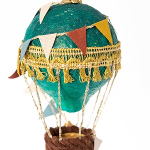 May include: A teal blue hot air balloon ornament with gold trim and colorful pennant flags. The balloon has a brown fabric basket and gold strings.