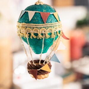 May include: A teal blue hot air balloon ornament with gold trim and colorful paper flags. The balloon has a brown wicker basket with more flags.