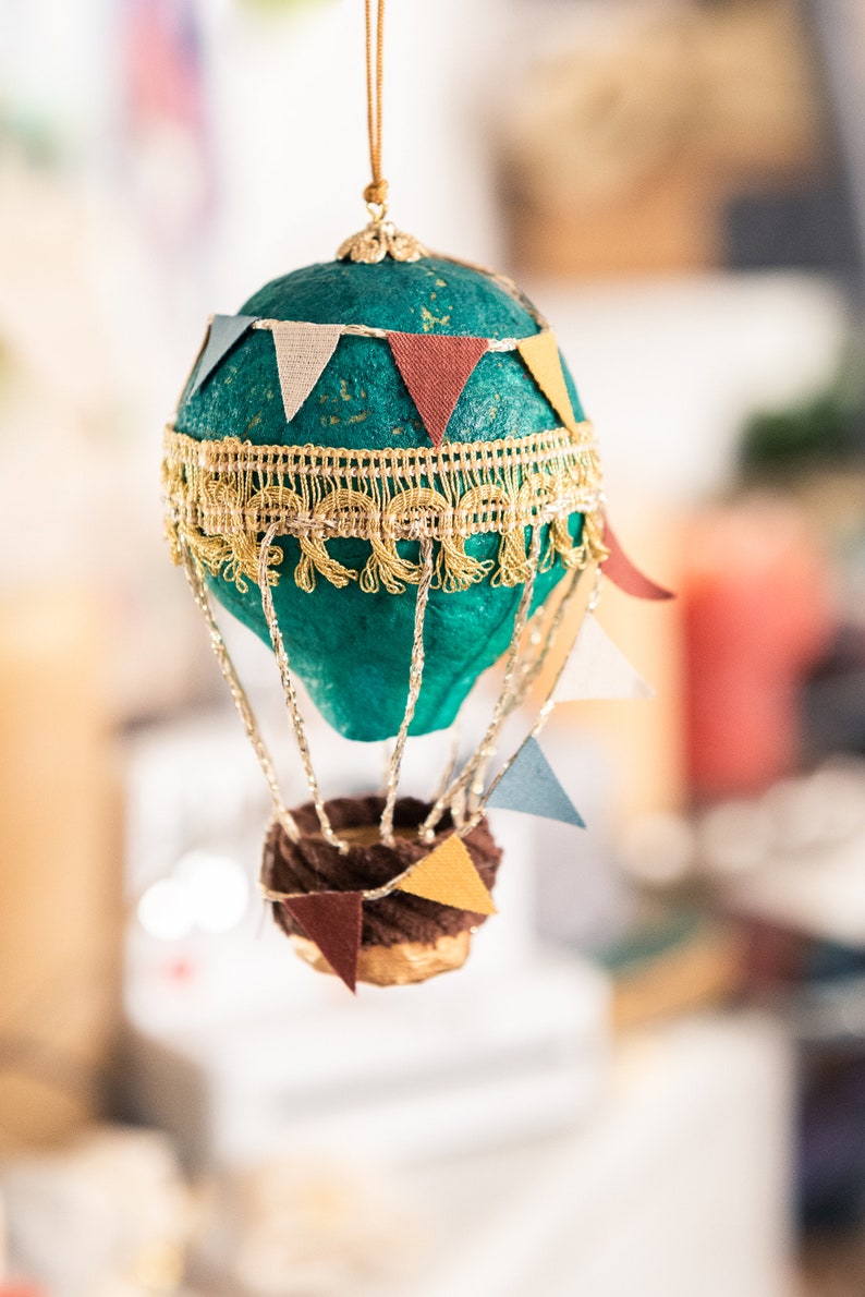 May include: A teal blue hot air balloon ornament with gold trim and colorful paper flags. The balloon has a brown wicker basket with more flags.