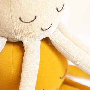 May include: Two plush octopus toys, one beige and one yellow, with embroidered eyes. The beige octopus is on top of the yellow octopus.