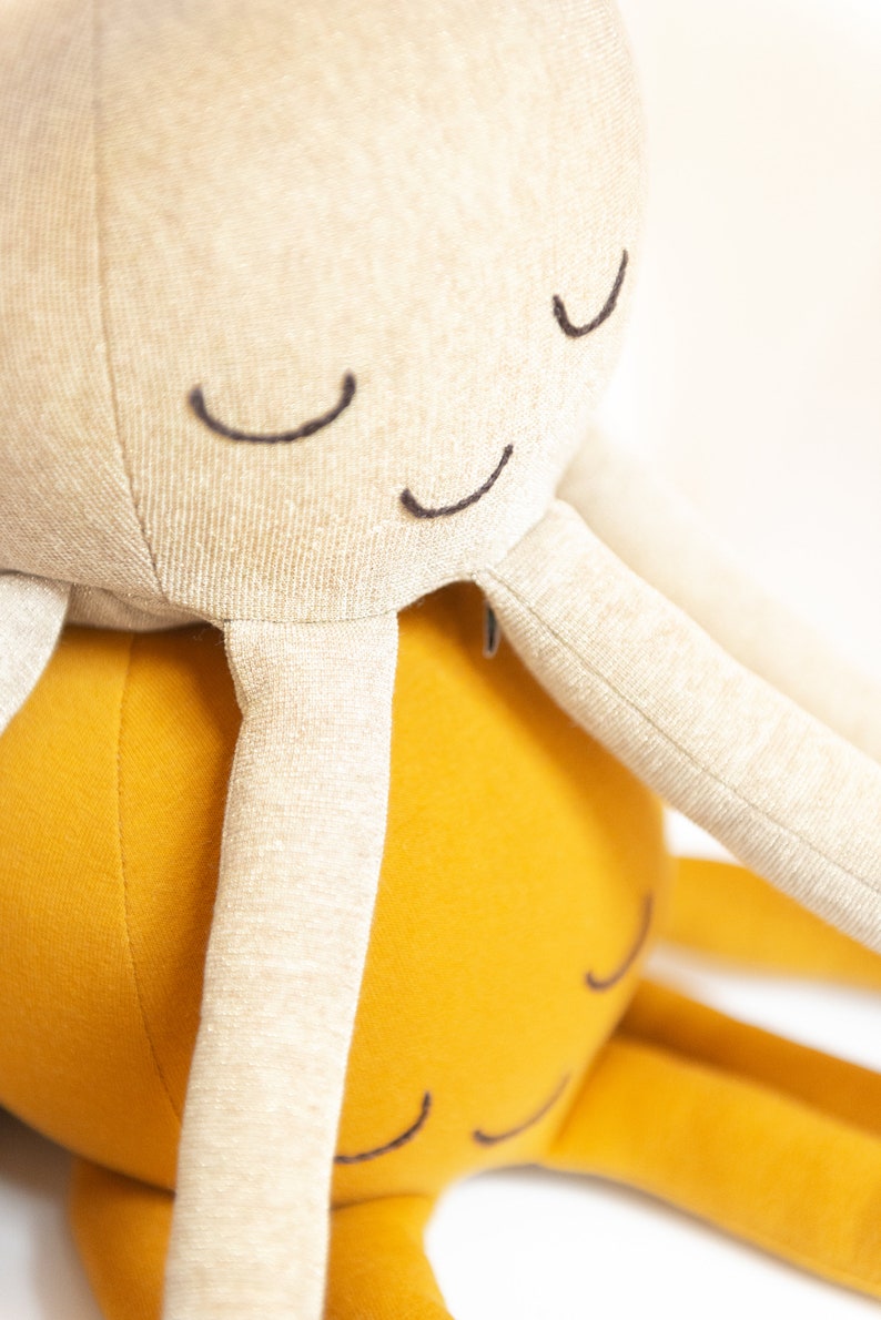 May include: Two plush octopus toys, one beige and one yellow, with embroidered eyes. The beige octopus is on top of the yellow octopus.