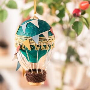 May include: A teal blue hot air balloon ornament with a gold trim and colorful pennant flags. The balloon has a brown wicker basket and is hanging from a gold string.