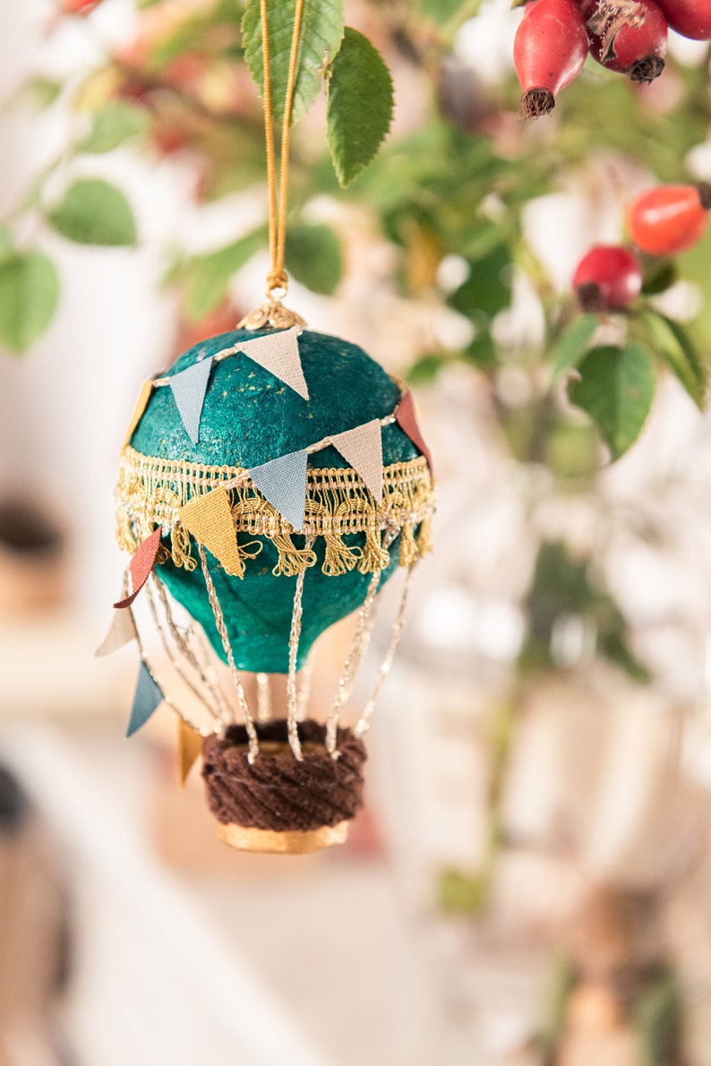 May include: A teal blue hot air balloon ornament with a gold trim and colorful pennant flags. The balloon has a brown wicker basket and is hanging from a gold string.