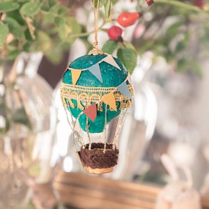 May include: A teal blue hot air balloon ornament with gold trim and colorful pennant flags. The balloon has a brown fabric basket with gold accents.