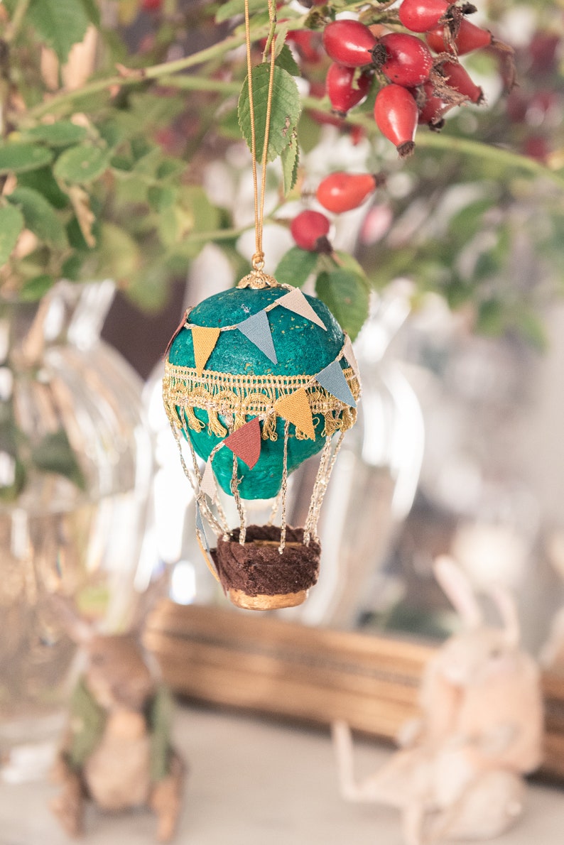 May include: A teal blue hot air balloon ornament with gold trim and colorful pennant flags. The balloon has a brown fabric basket with gold accents.