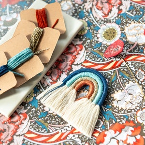 Puede incluir: Un tapiz de macramé de arcoíris con borlas blancas. El macramé está hecho con hilo azul, verde, naranja y dorado. El macramé está sobre un fondo de tela estampada.