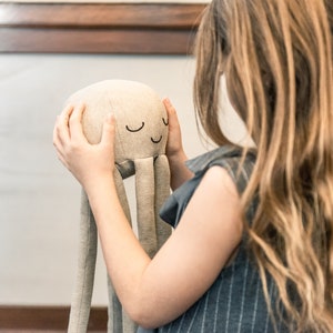 May include: A young person is holding a soft, grey, plush octopus toy with embroidered eyes and a smiling mouth. The toy has long, thin arms.