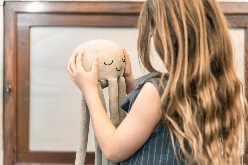 May include: A young person is holding a soft, grey, plush octopus toy with embroidered eyes and a smiling mouth. The toy has long, thin arms.