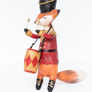 Può includere: Una statuetta di volpe in cartapesta fatta a mano vestita da tamburino. La volpe indossa un'uniforme rosso e nero con rifiniture dorate, un cappello nero con una catena dorata e stivali neri. La volpe tiene due bacchette e ha un tamburo rosso e dorato legato alla schiena.
