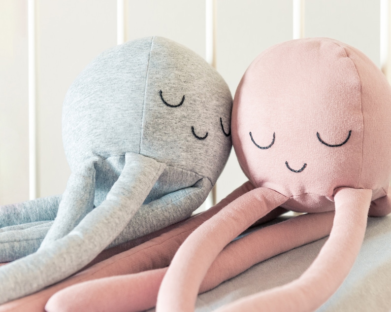 May include: Two plush octopus toys with embroidered eyes and smiles. One octopus is grey and the other is pink.