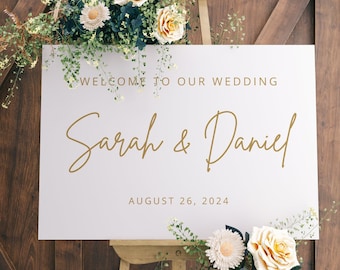 May include: White wedding sign with gold lettering on an easel. The sign reads "Welcome to our wedding Sarah & Daniel August 26, 2024". The sign is decorated with white and yellow flowers.
