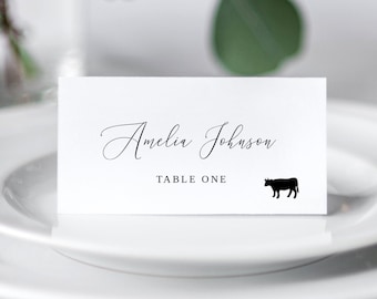 May include: White place card with black text that reads "Amelia Johnson" and "Table One". A black silhouette of a cow is to the right of the text.