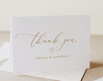 Puede incluir: Una tarjeta de agradecimiento blanca con letras doradas que dice "Gracias" con un pequeño símbolo de corazón y los nombres "Emilia & Jonathan" debajo.