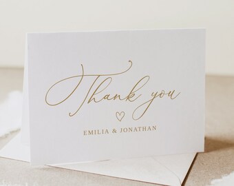 Puede incluir: Una tarjeta de agradecimiento blanca con letras doradas que dice "Gracias" con un pequeño símbolo de corazón y los nombres "Emilia & Jonathan" debajo.
