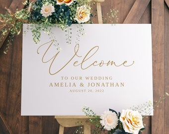May include: White wedding welcome sign with gold calligraphy lettering. The sign reads "Welcome to our wedding Amelia & Jonathan August 20, 2022". The sign is propped up on an easel with flowers in front of it.