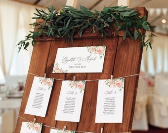 May include: A rustic wooden wedding seating chart with floral accents. The chart features a sign with the names "Elizabeth & Michael" and "Find Your Seat." Individual cards are clipped to twine, each with guest names and table numbers.