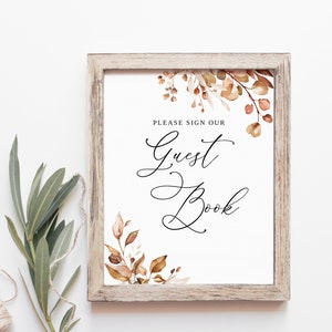 May include: A rustic wooden frame with a white background and a sign that says "Please sign our Guest Book". The sign is decorated with a watercolor illustration of fall leaves in shades of orange, brown, and beige.