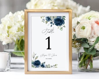 May include: A gold-framed table number card with a watercolor floral design in shades of blue and green. The card reads "Table 1" and includes the names "Harper & James" and the date "08.14.2022".