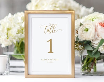 Könnte beinhalten: Goldene Tischkartenhalter mit der Aufschrift "Tisch 1" in goldener Schrift. Die Karte enthält auch die Namen "Sarah & Michael" und das Datum "06.14.2022".
