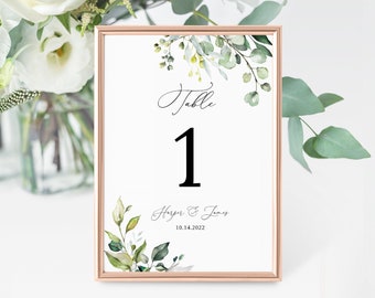 May include: A white table number card with a gold frame. The card has a green floral design and the text "Table 1 Harper & James 10.14.2022".