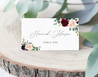 May include: White wedding place card with a floral design in shades of burgundy, pink, and blue. The card reads "Hannah Johnson" and "Table One".