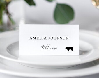 May include: White place card with black text that reads "Amelia Johnson, table one". A small black silhouette of a cow is to the right of the text.