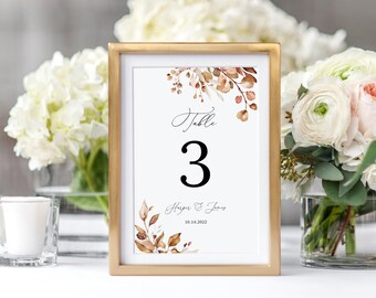 May include: A gold-framed table number card with a white background and brown floral illustrations. The card reads "Table 3" and "Harper & James 10.14.2022".