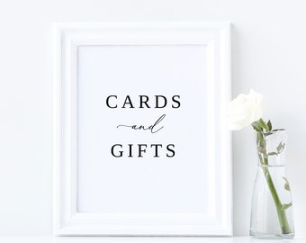 May include: White framed sign with black text that reads "CARDS and GIFTS" with a single white rose in a clear glass vase on a white surface.