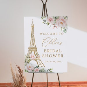 May include: A white bridal shower welcome sign with a gold Eiffel Tower illustration and pink floral accents. The text reads "Welcome to Chloe's Bridal Shower August 16, 2025".