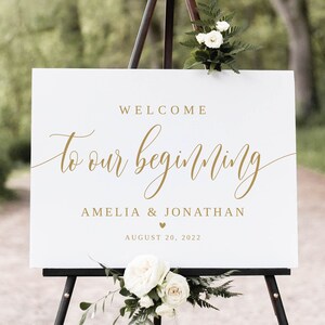 May include: White wedding welcome sign with gold lettering that reads "Welcome to our beginning Amelia & Jonathan August 20, 2022". The sign is on a black easel with white flowers.