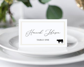 Op de afbeelding: Witte plaatskaart met een gouden rand en een zwart silhouet van een koe. De tekst op de kaart luidt "Hannah Johnson, Tafel Eén."